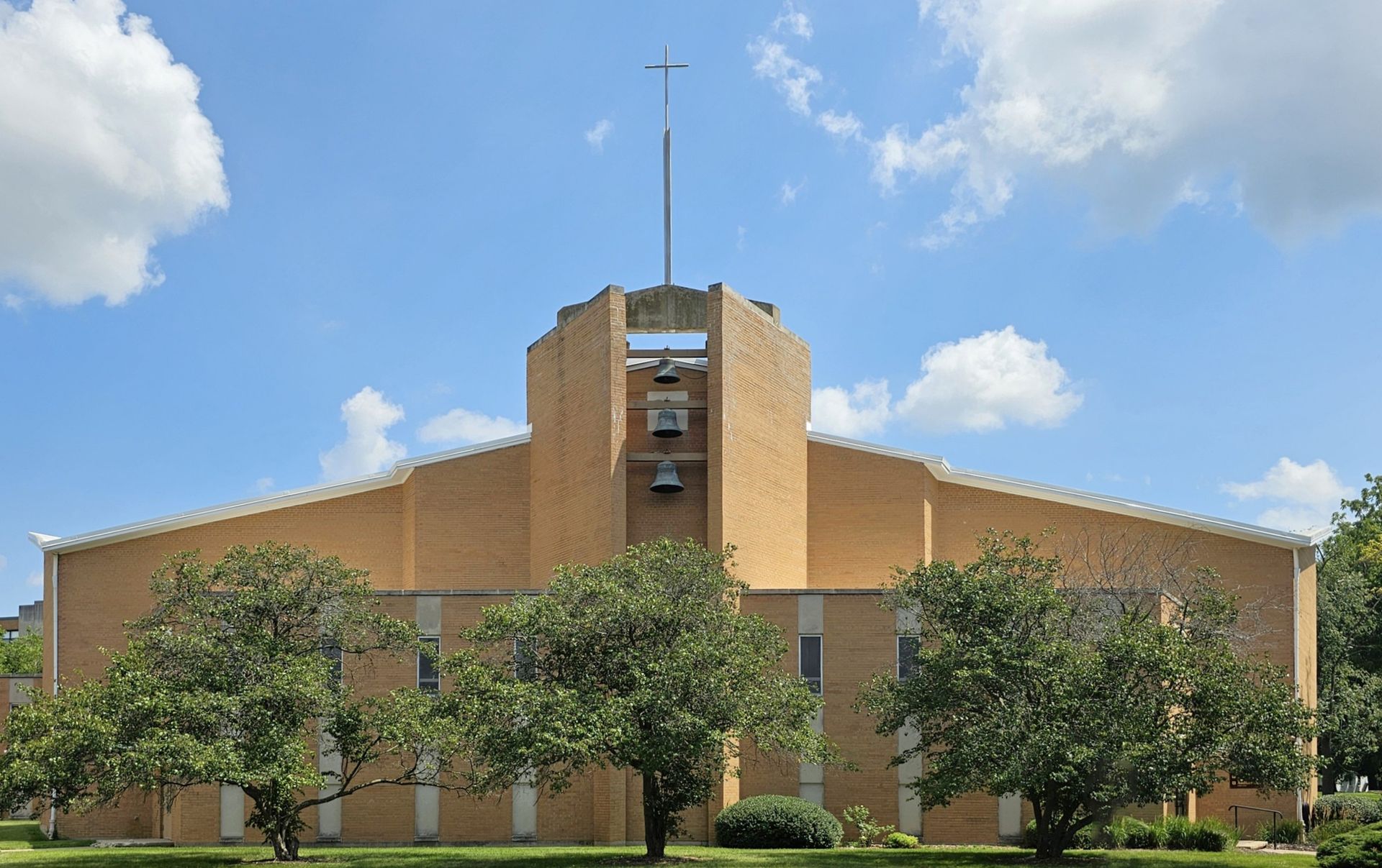 A tan brick church building under a bright blue sky with scattered clouds, featuring a central bell tower and a cross.