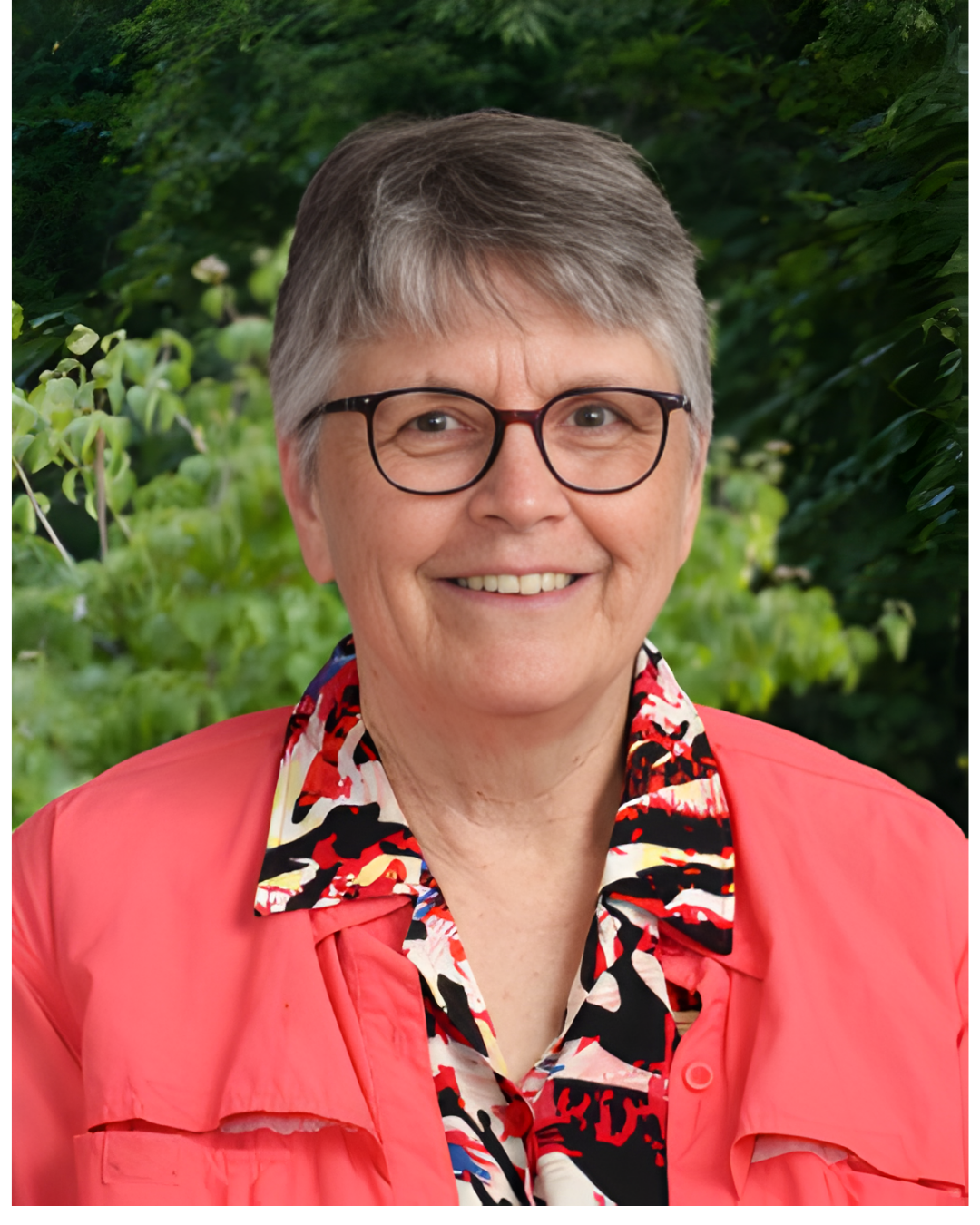 A person with short, grey hair and glasses wearing a coral jacket over a colorful patterned shirt against a leafy background.