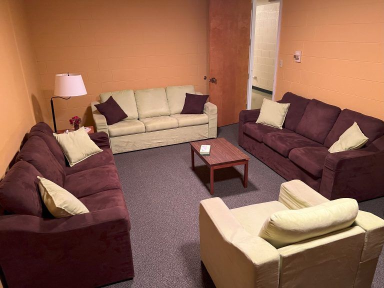 A room with two dark purple sofas, a cream sofa, a matching armchair, a coffee table, and a floor lamp on orange walls.