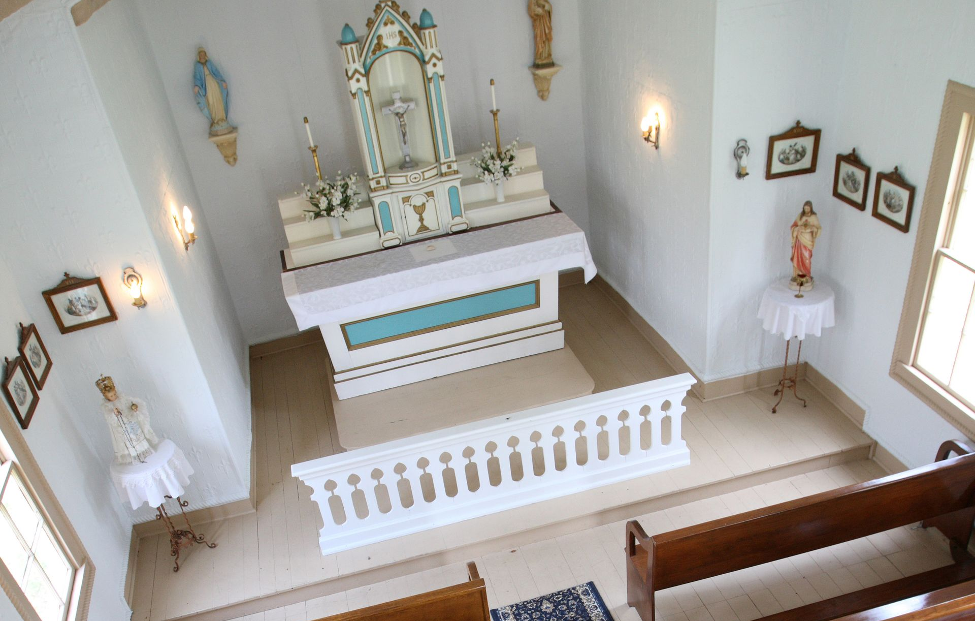 A high-angle view of a small chapel interior featuring a white altar, pews, religious artwork, and pedestals.