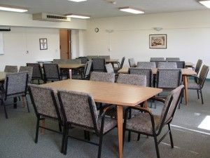 An empty meeting room with rows of wooden tables and gray upholstered chairs arranged on a gray carpet.