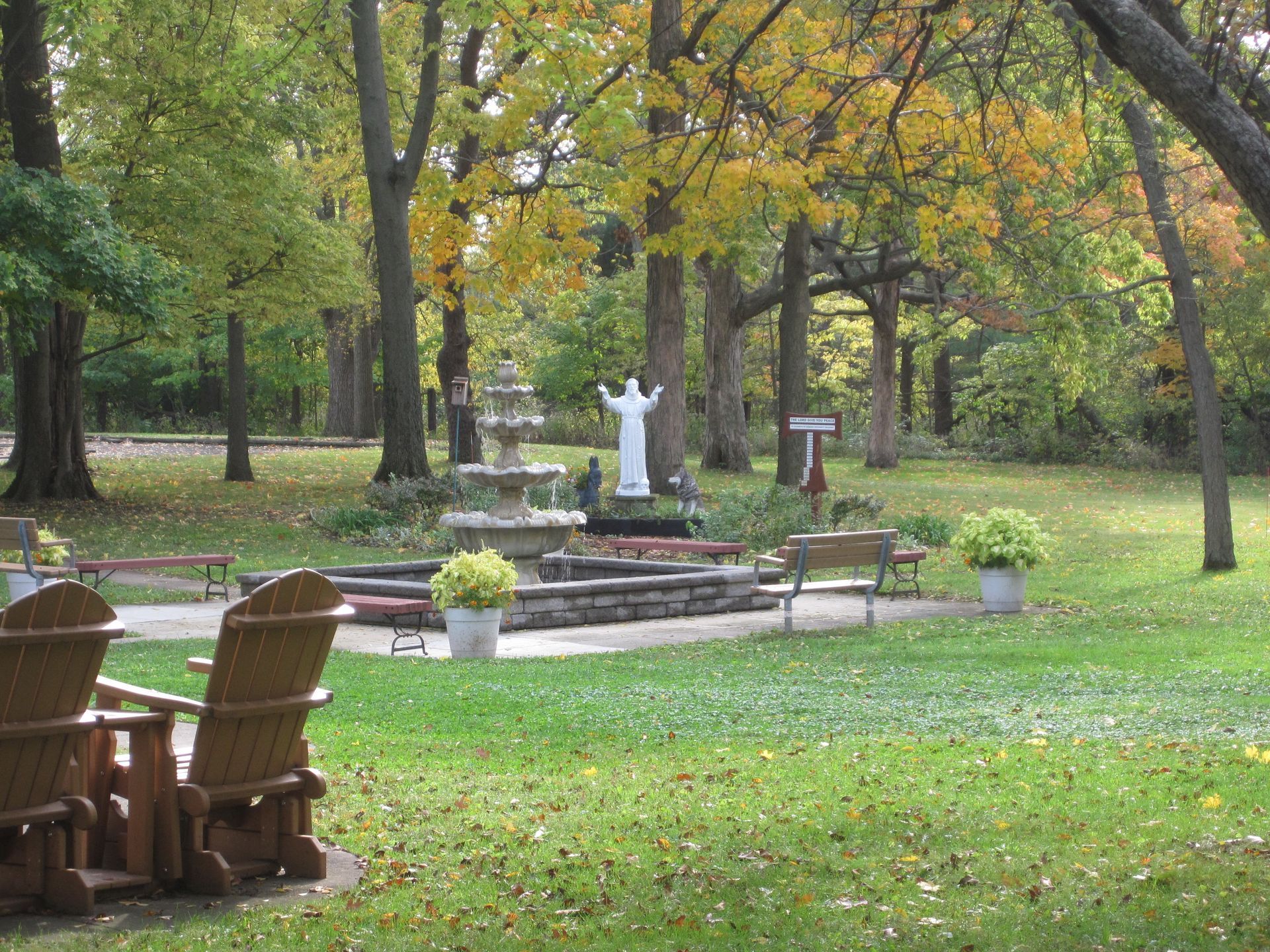 A stone fountain and a white statue of an angel in a grassy, tree-filled park with wooden chairs in the foreground.