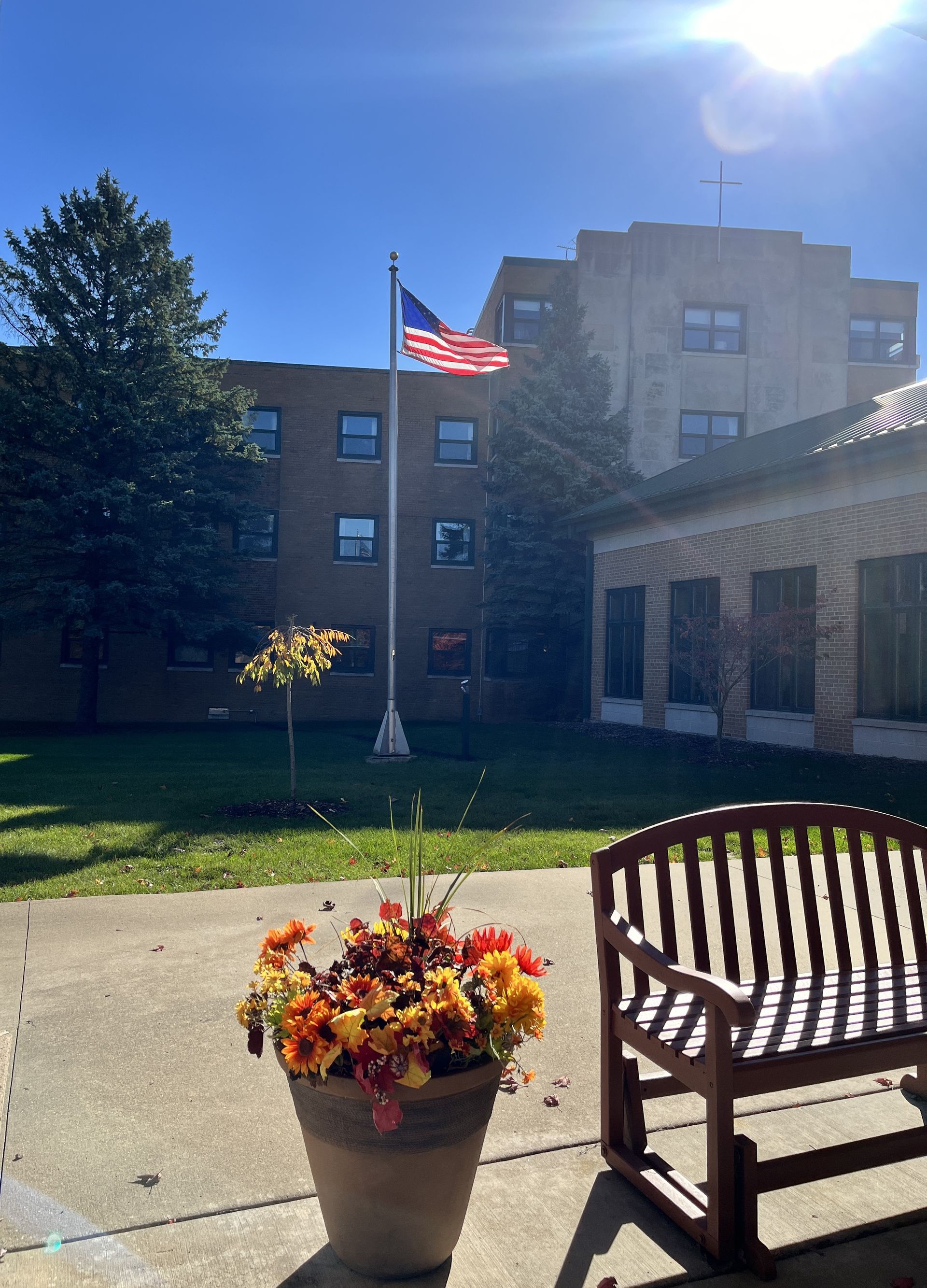 A wooden bench sits next to a potted plant, with a courtyard, flag, and brick building in the background.