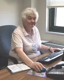 A person with short light hair sits at a desk in an office setting, typing on a keyboard and using a computer mouse.