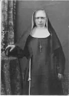 A nun in a black habit and white wimple stands beside a small table with a crucifix, looking toward the camera.