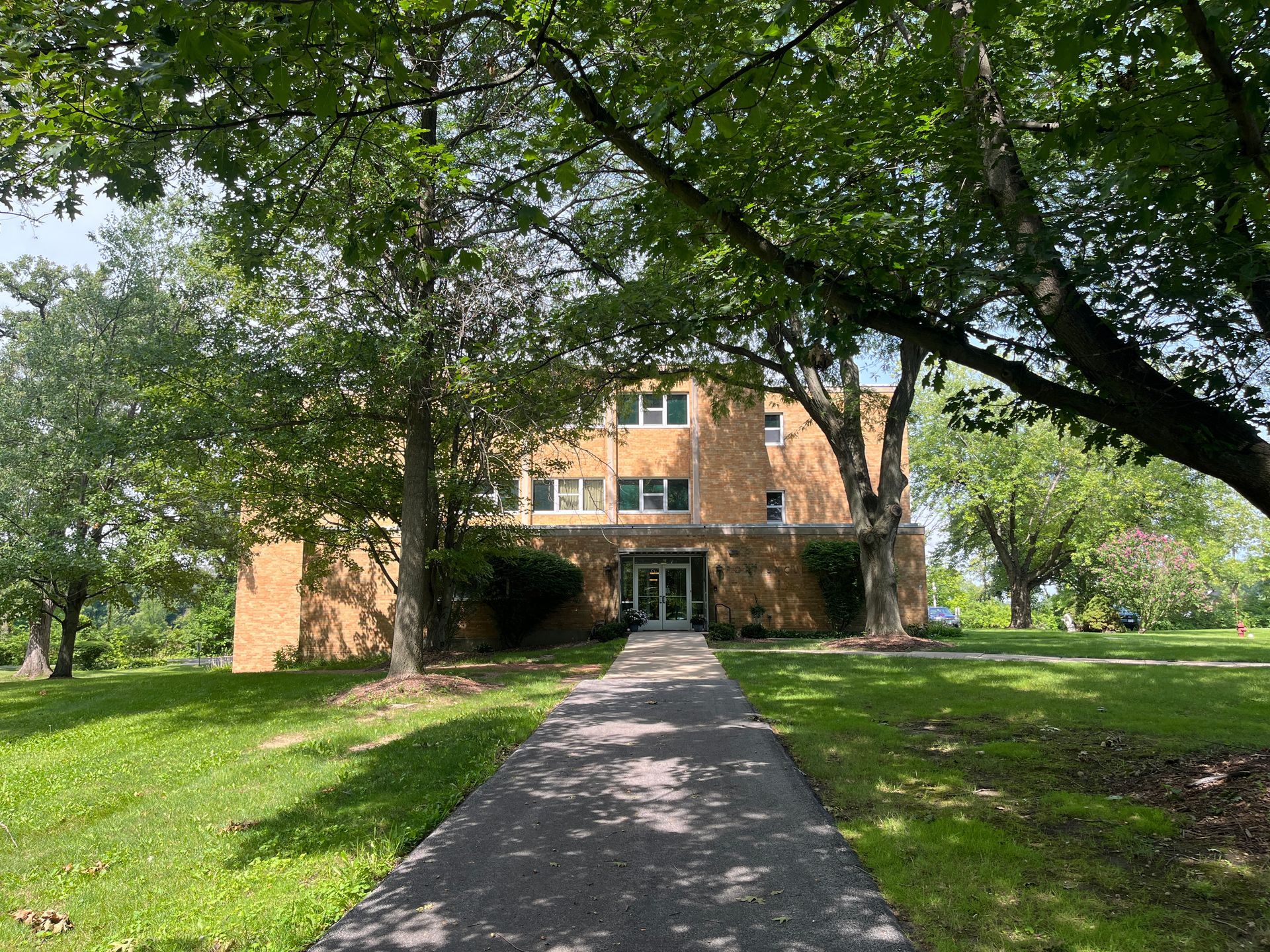 A tan, three-story brick building set behind lush green trees, with a concrete path leading to the entrance.