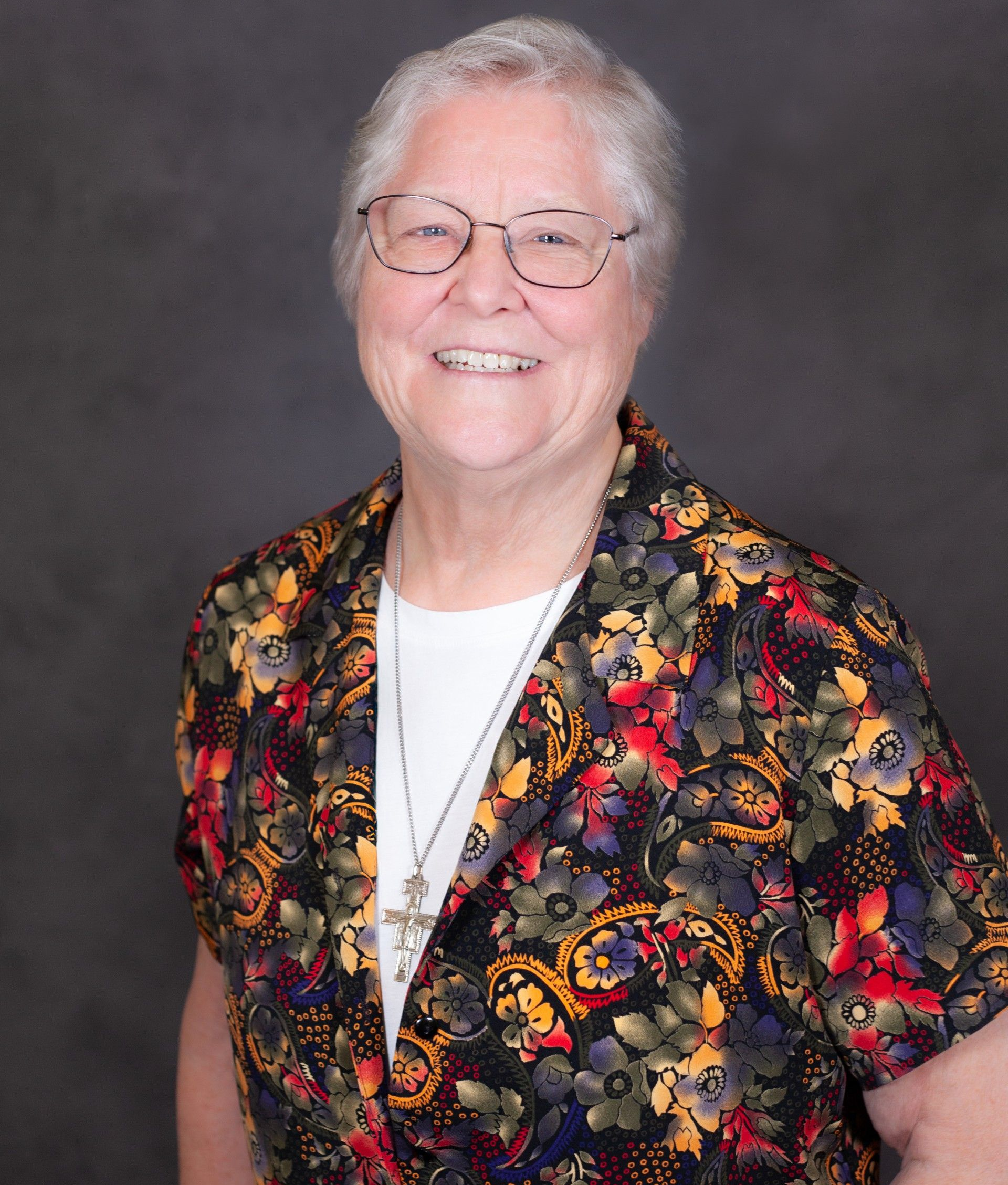 An older person smiling in a floral-patterned blouse and a cross necklace against a gray background.