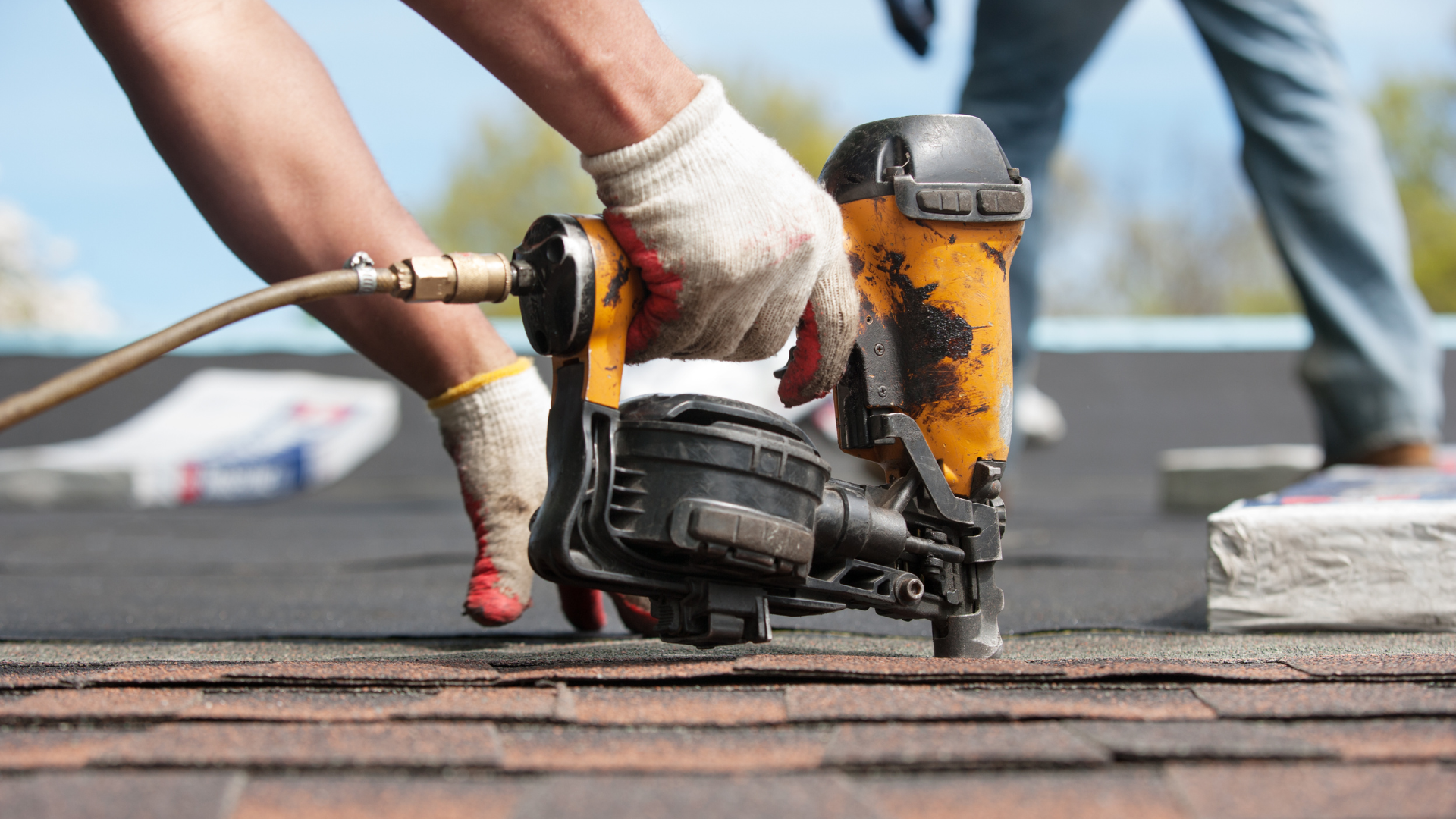 Gloved hands use a nail gun to fasten roofing shingles on a rooftop