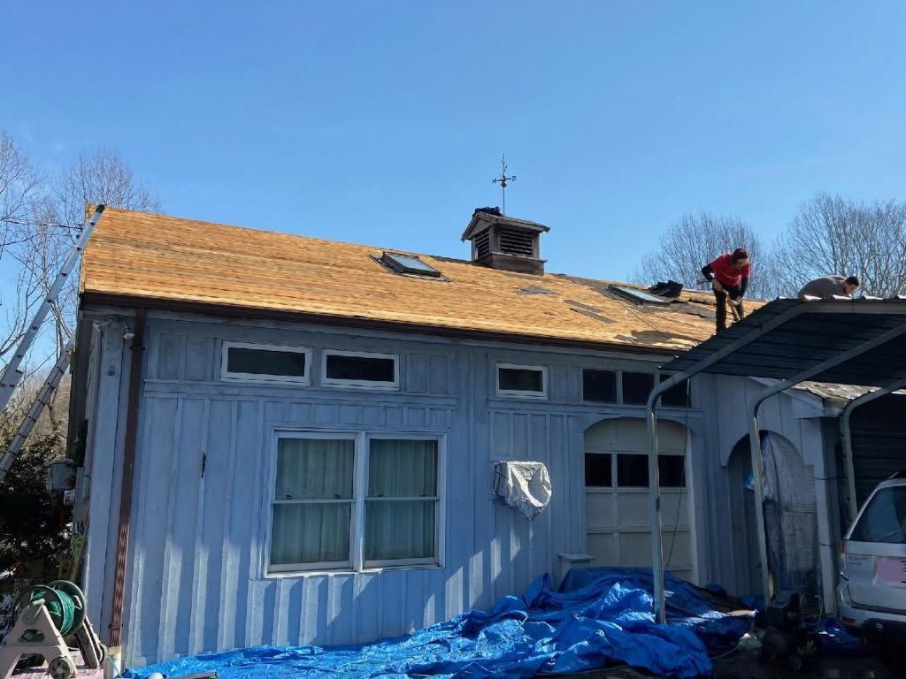 Damaged house exterior with torn roof, blue tarps, and debris in the yard under a clear sky