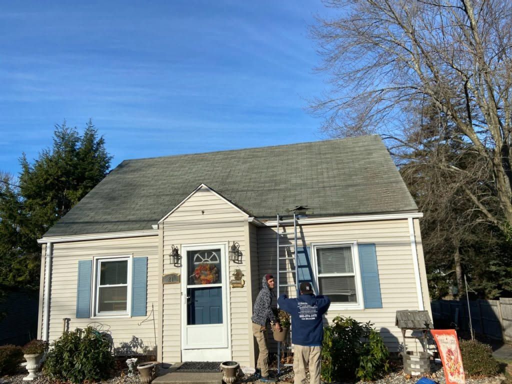 Small cream house with blue shutters; two people stand on the front stoop under a ladder.