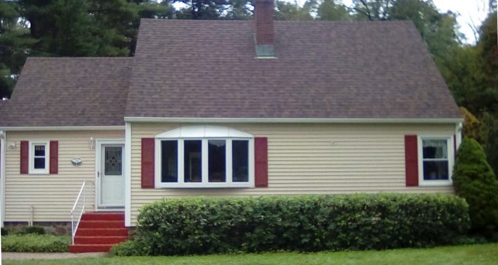 Small beige house with red shutters and steps, white-trimmed bay window, and a neatly trimmed hedge.
