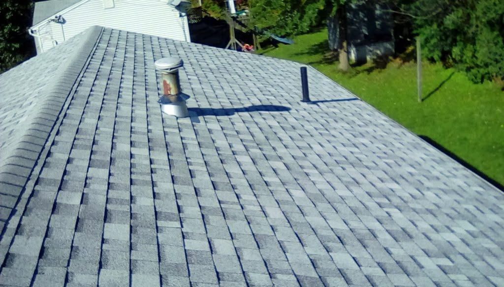 Gray shingle roof with two vents and a shadowed chimney, bordered by green trees and lawn