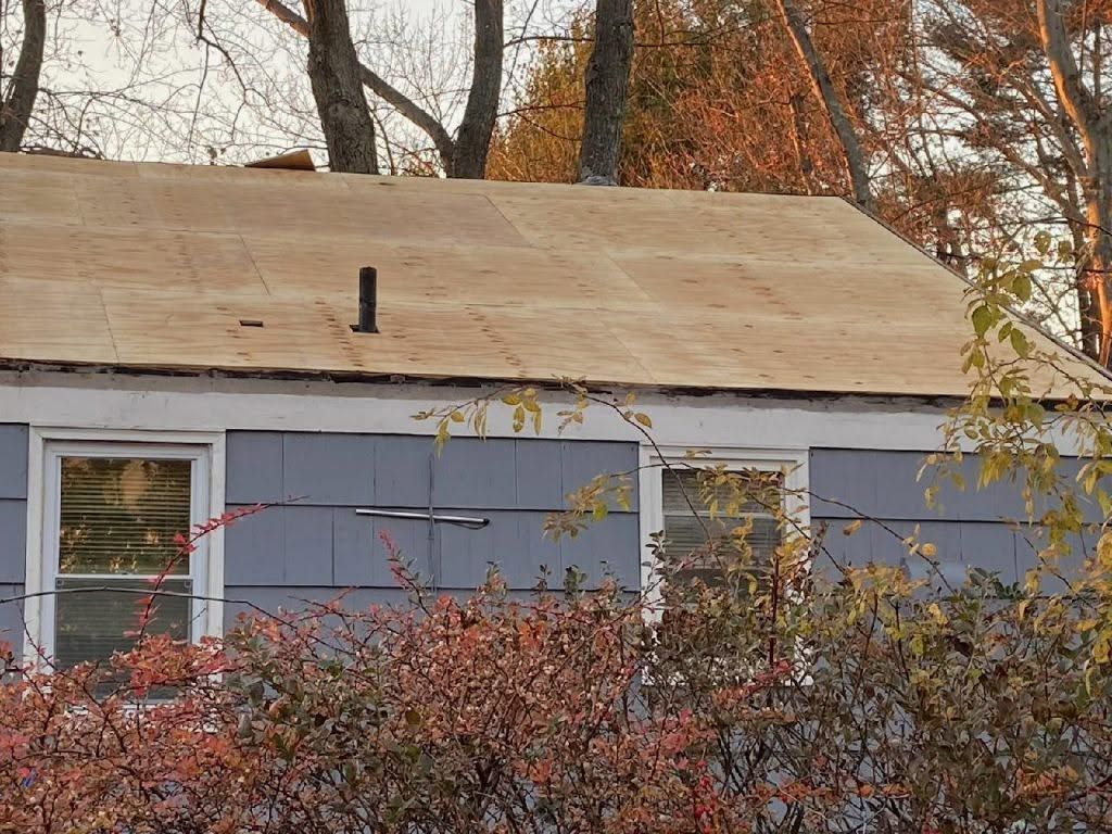 Blue house with a partially replaced plywood roof amid autumn trees and bushes