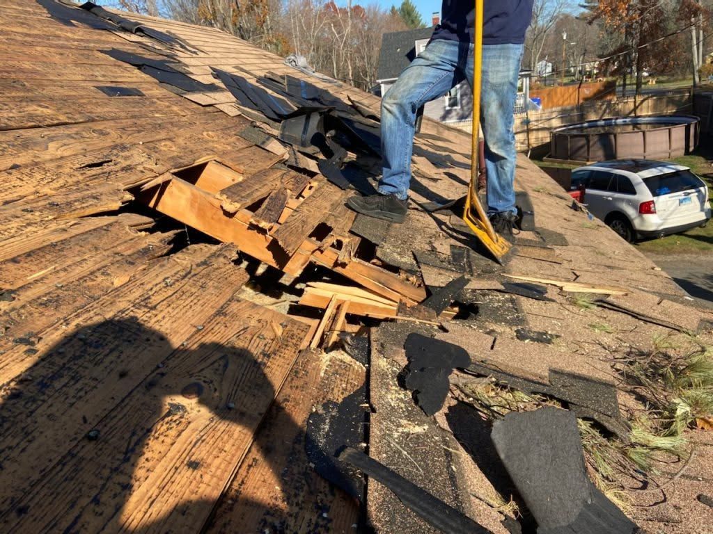 Worker repairing damaged shingle roof with exposed underlayment and scattered debris near a ladder.