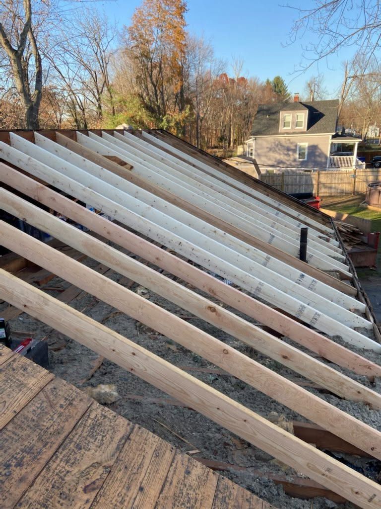 Roof framing with exposed white rafters under construction beside a wooded suburban yard.