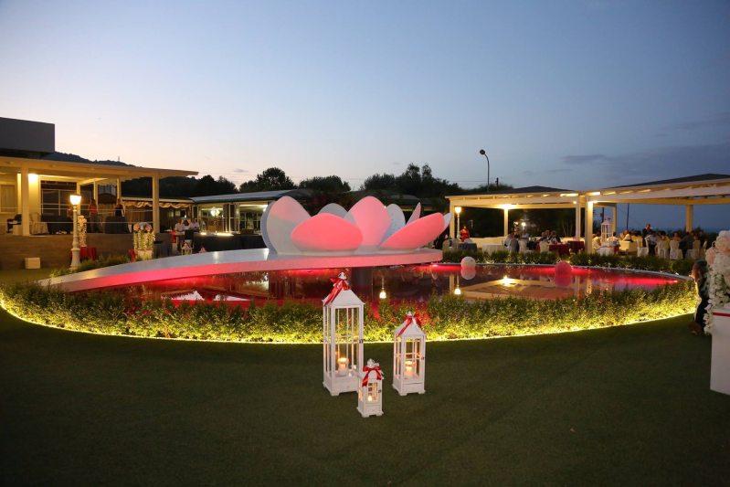 Giardino notturno con una fontana di fiori rosa luminosi e lanterne appese su un prato.