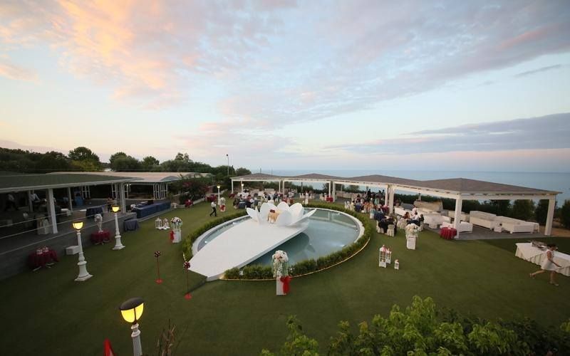 Ricevimento di nozze all'aperto a bordo piscina al tramonto, con gli ospiti disposti su un prato sotto tendoni bianchi e una terrazza coperta.