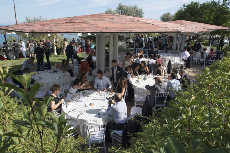 Persone sedute a tavoli rotondi sotto un gazebo ombreggiato all'aperto, intente a cenare insieme.