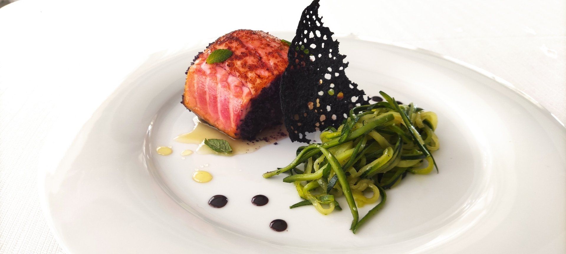 Filetto di pesce rosa scottato con verdure e salsa su un piatto bianco, guarnito con una cialda nera croccante.