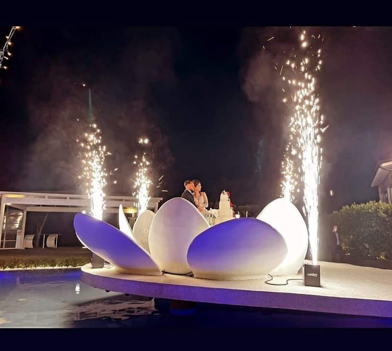 Scena di matrimonio notturna con sculture di fiori bianchi illuminate, fuochi d'artificio e una coppia su un palco a bordo piscina.
