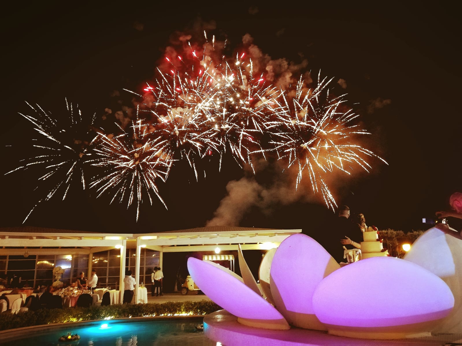 Fuochi d'artificio esplodono sopra la piscina di un resort notturno, con una fontana scultorea di un viola intenso in primo piano.