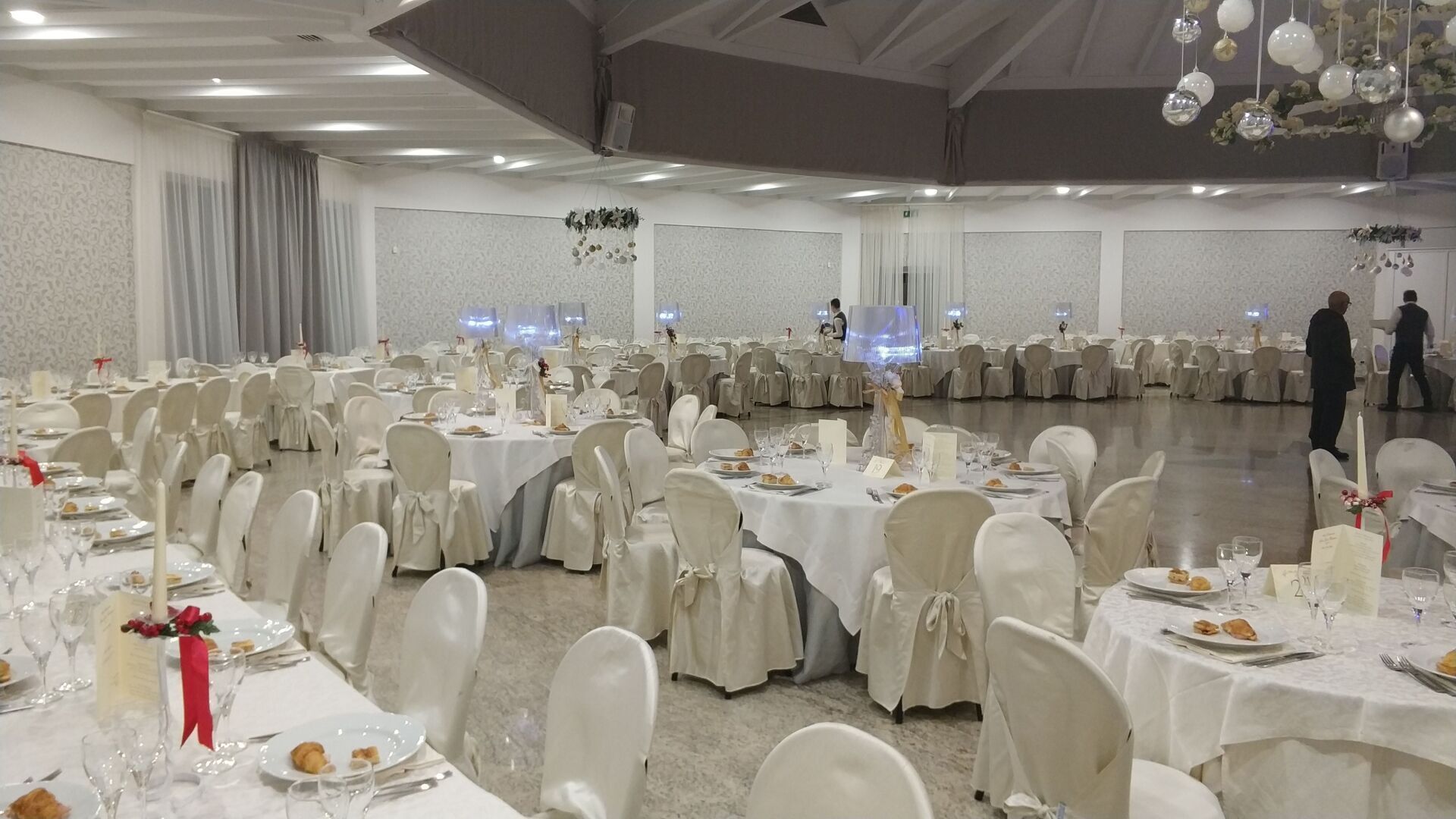 Elegante sala per banchetti con numerosi tavoli rotondi, coprisedie bianchi, lampadari e alcuni ospiti sullo sfondo.