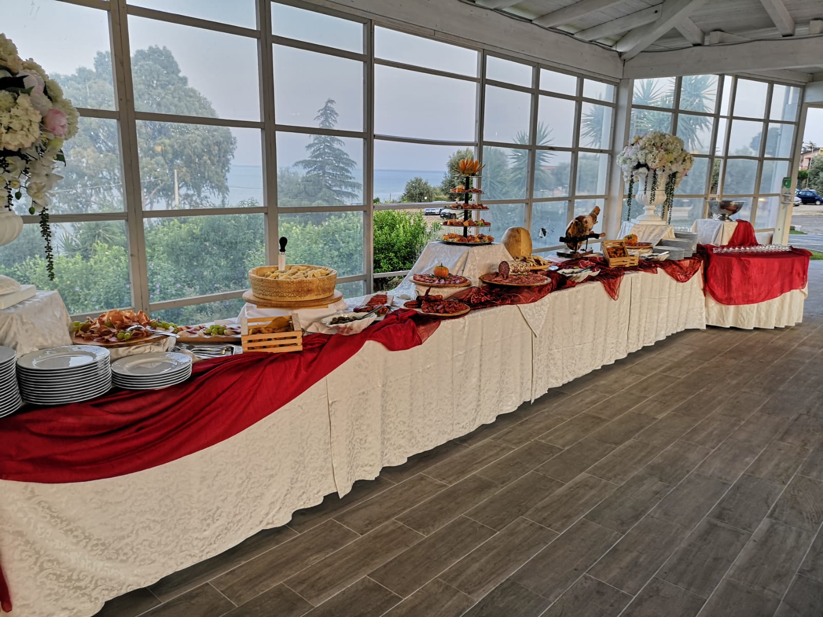 Un tavolo da buffet con drappeggi rossi e bianchi è allestito in una sala da pranzo con pareti a vetri che si affaccia sul verde.