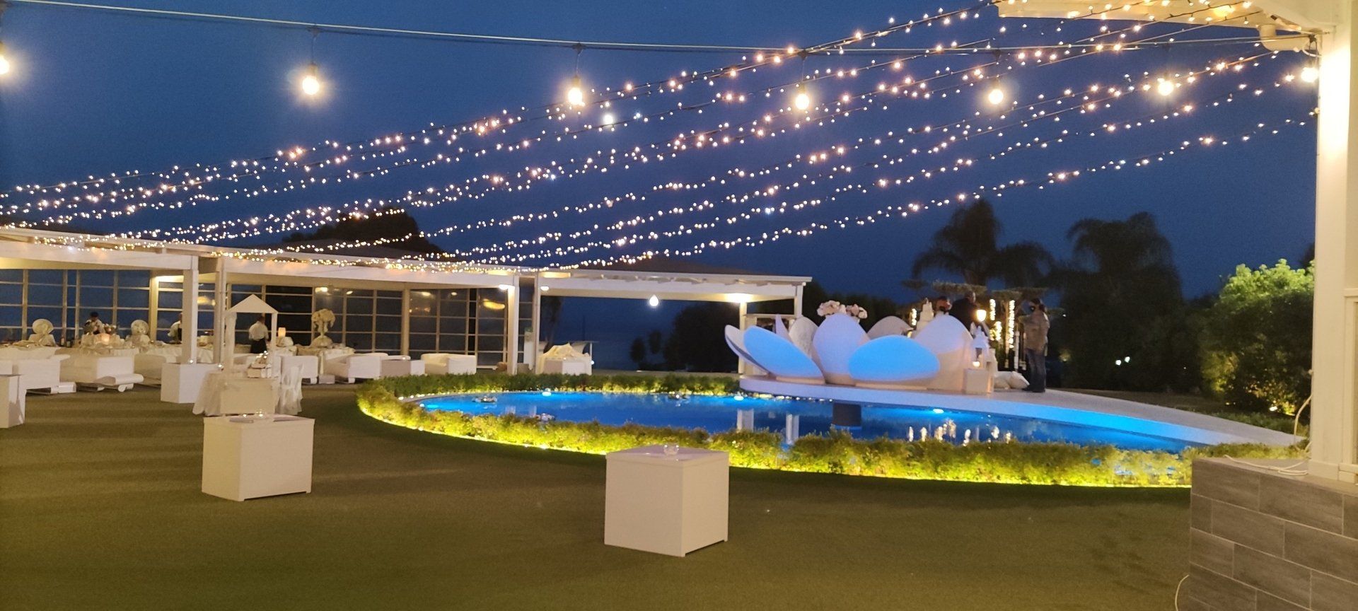 Patio esterno serale con luci decorative, sedie a sdraio e una fontana illuminata sotto un cielo blu scuro