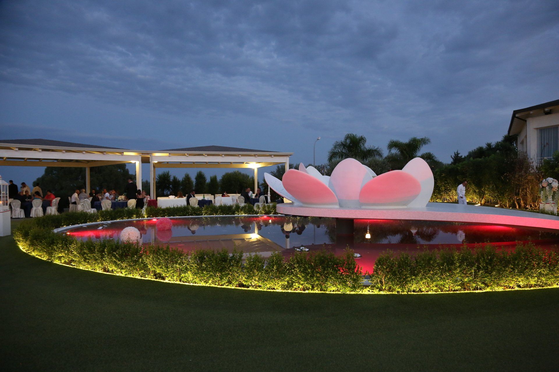 Fontana da giardino notturna con scultura a forma di loto rosa luminosa e riflessi in uno stagno circolare.
