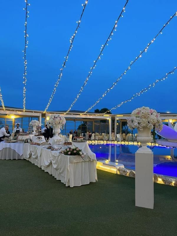 Banchetto serale all'aperto con tavoli apparecchiati con drappeggi bianchi, centrotavola floreali e luci decorative a bordo piscina.