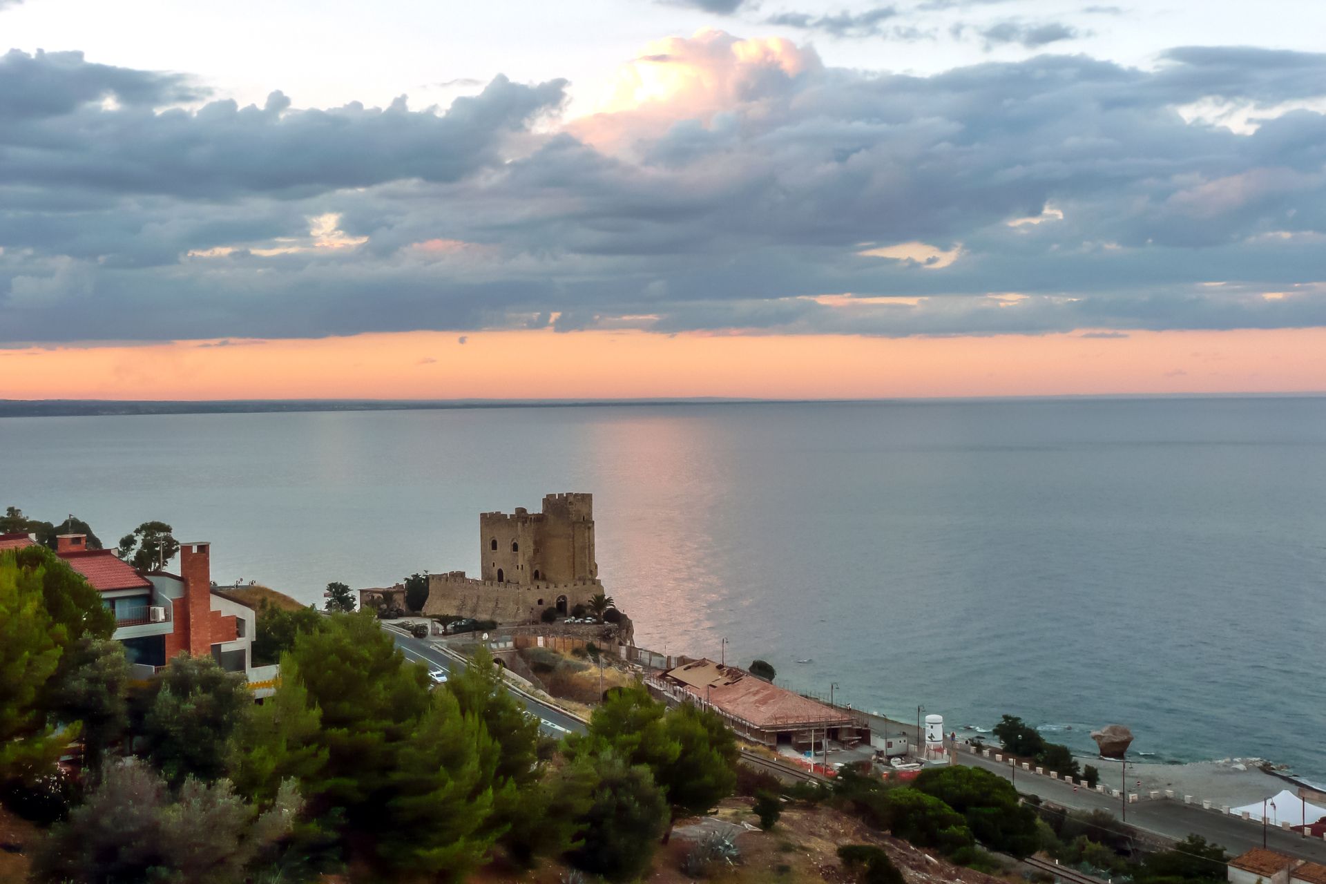 Cittadina costiera al tramonto con mare, cielo nuvoloso e un castello di pietra arroccato su una scogliera.
