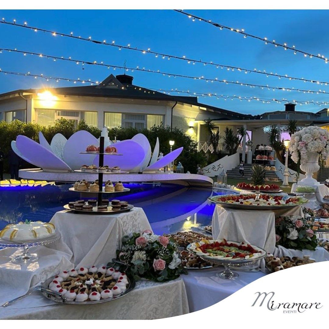 Elegante buffet all'aperto a bordo piscina, illuminato al crepuscolo, con decorazioni floreali bianche e luci a catena sospese.