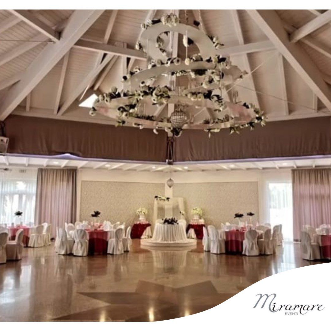 Elegante sala per ricevimenti di nozze con drappeggi bianchi, tavoli rotondi e centrotavola con lampadario floreale.