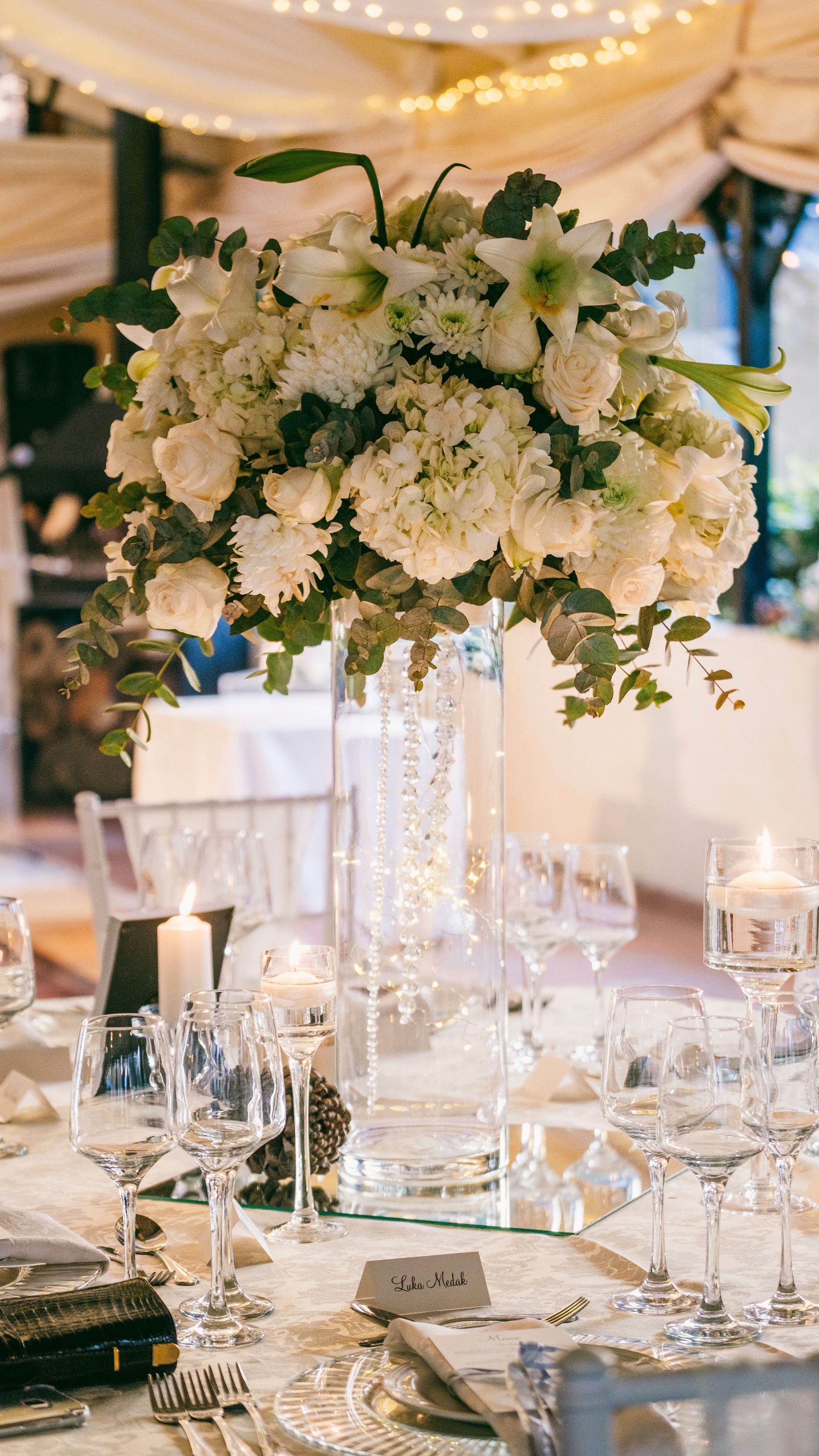 Elegante tavolo per il ricevimento di nozze con un alto centrotavola floreale bianco e bicchieri di cristallo sotto luci soffuse.