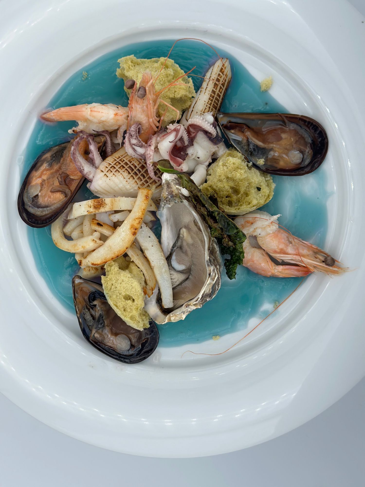 Assortimento di frutti di mare con cozze, gamberi, calamari e guarnizioni su un piatto bianco con salsa blu.
