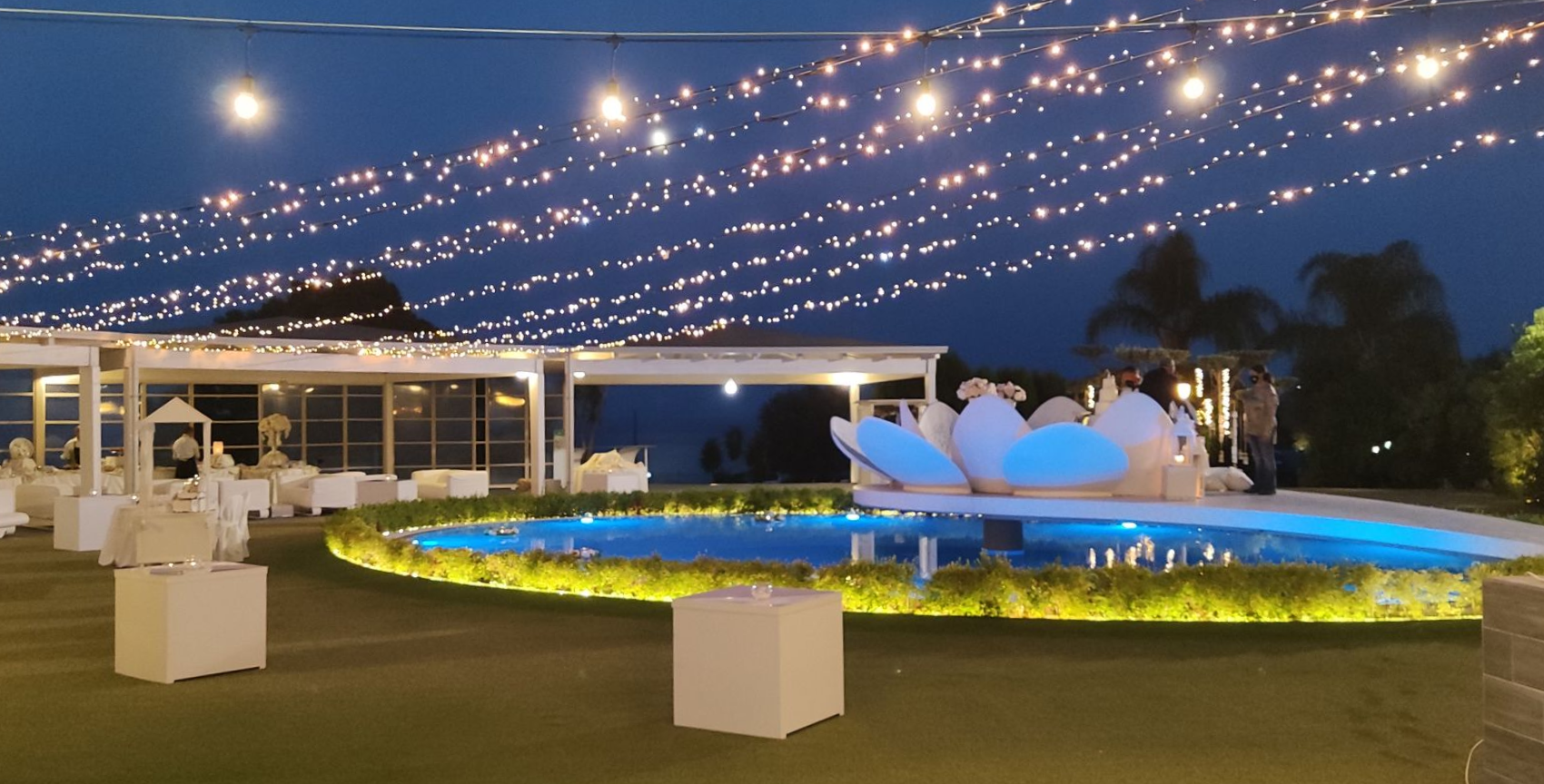 Spazio per eventi all'aperto al crepuscolo con luci decorative, sedute bianche e una fontana illuminata.