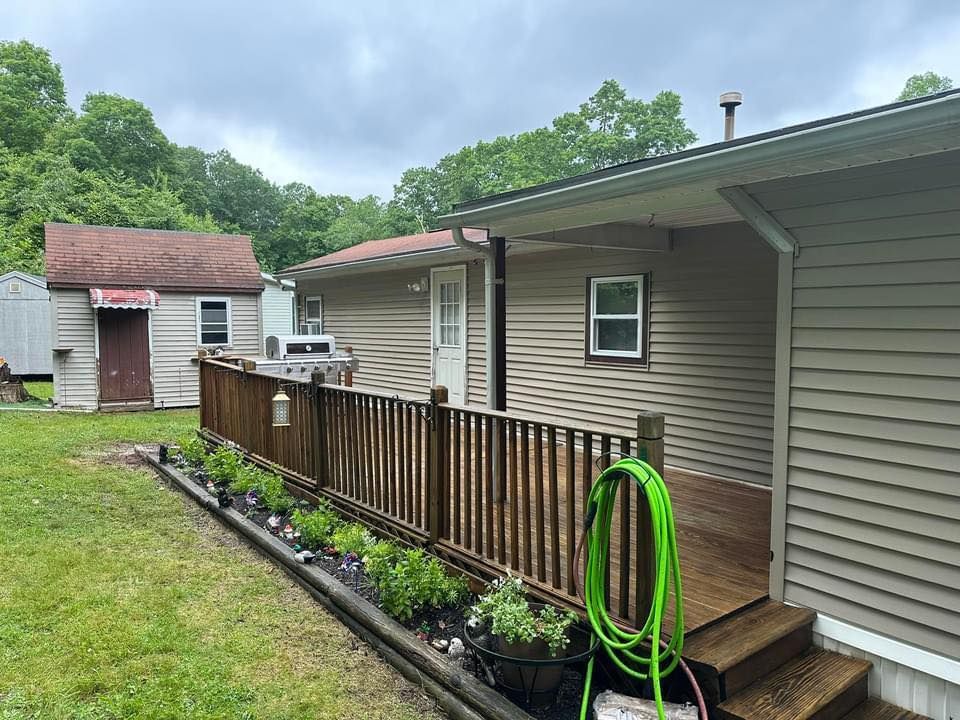 Exterior view of a house with a wooden deck and fence, green hose, small shed, and trees.