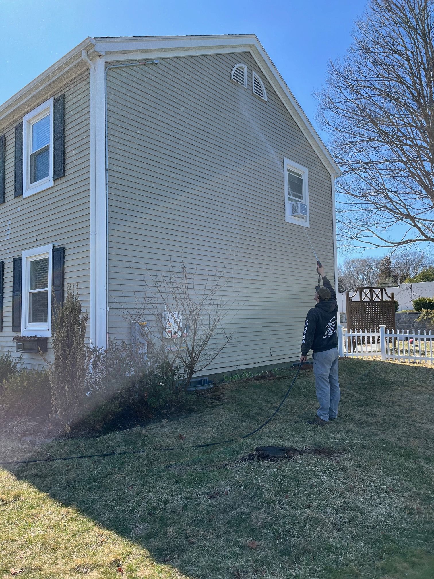 Person pressure washing the side of a two-story beige house on a sunny day.
