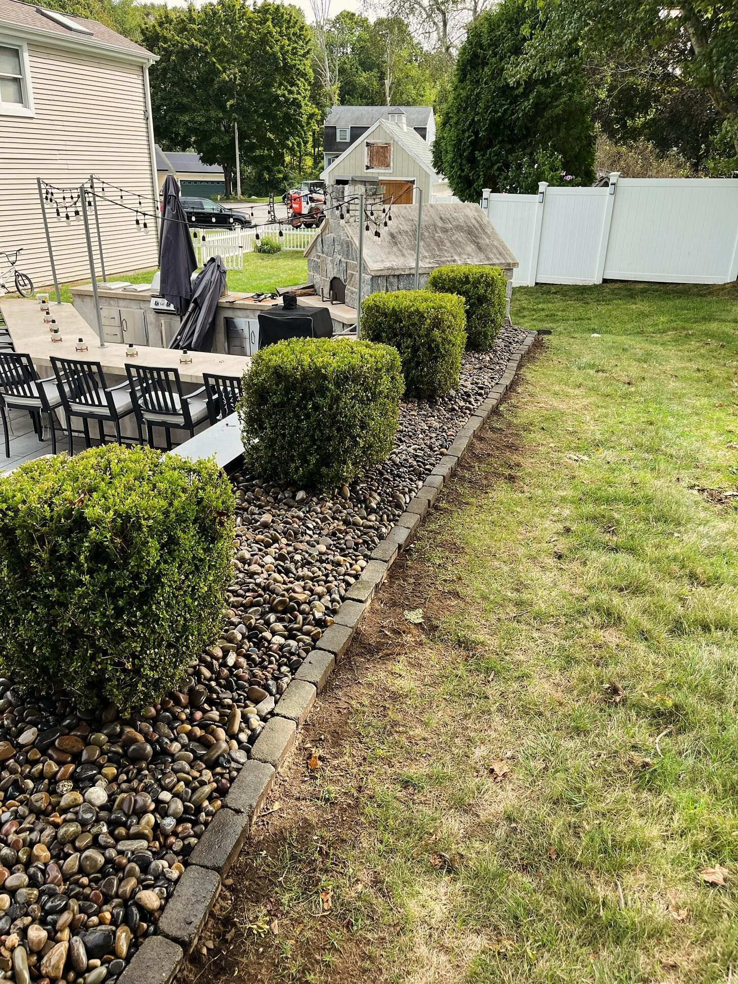 A person digs a trench in a backyard near a small excavator, a tree, and a house.