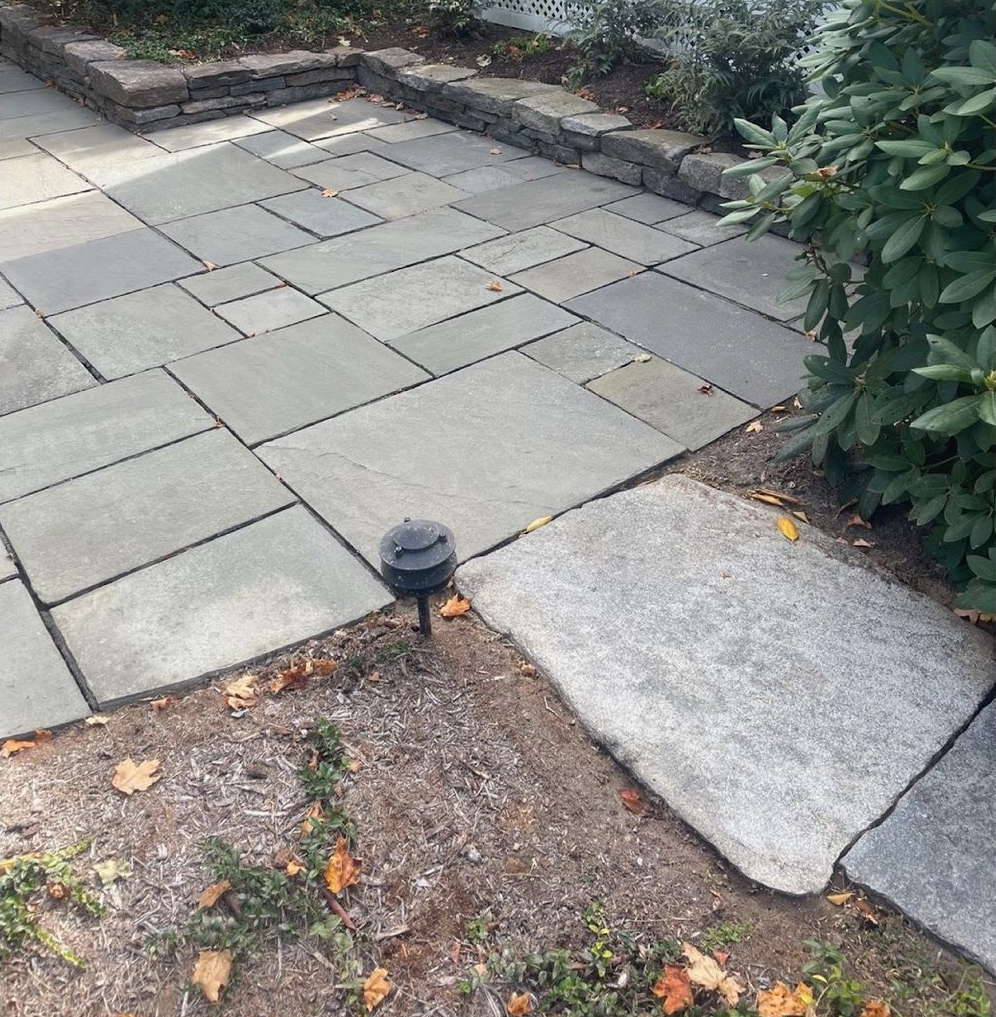 Stone patio with a low retaining wall. A light fixture and a stone pathway are in the foreground.