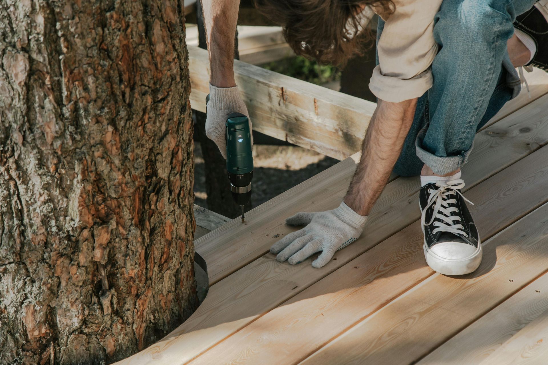 Person wearing work gloves using a drill to install wooden deck boards around a tree.