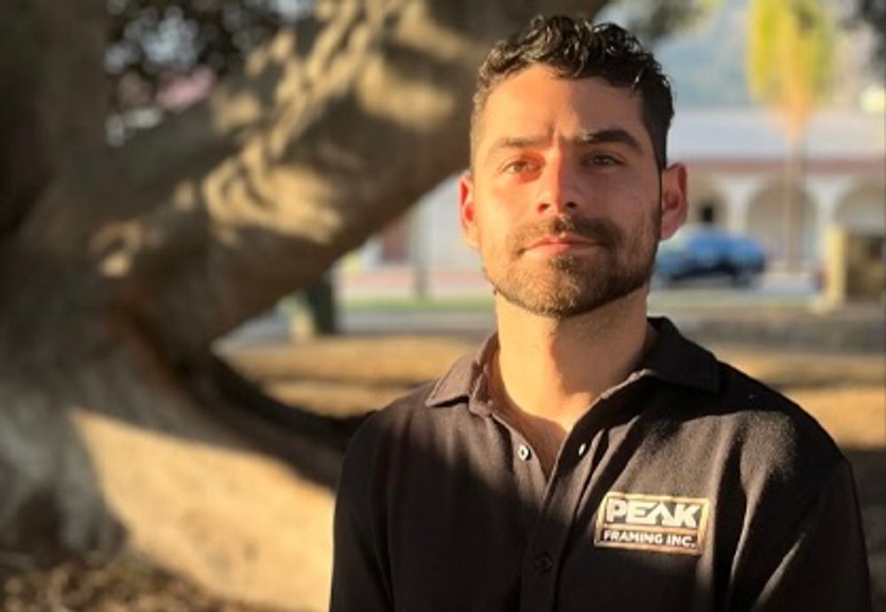 Man with a beard in a black polo shirt, standing in front of a tree outdoors, looking at the camera.
