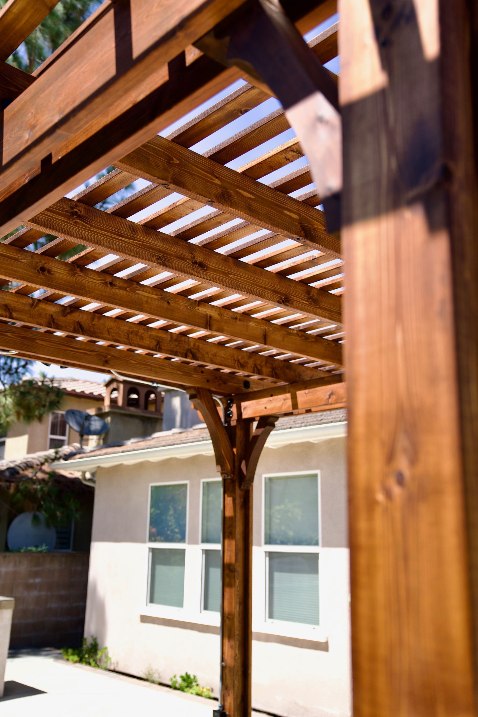 Wooden pergola with slatted roof over a patio beside a house with white-trimmed windows.