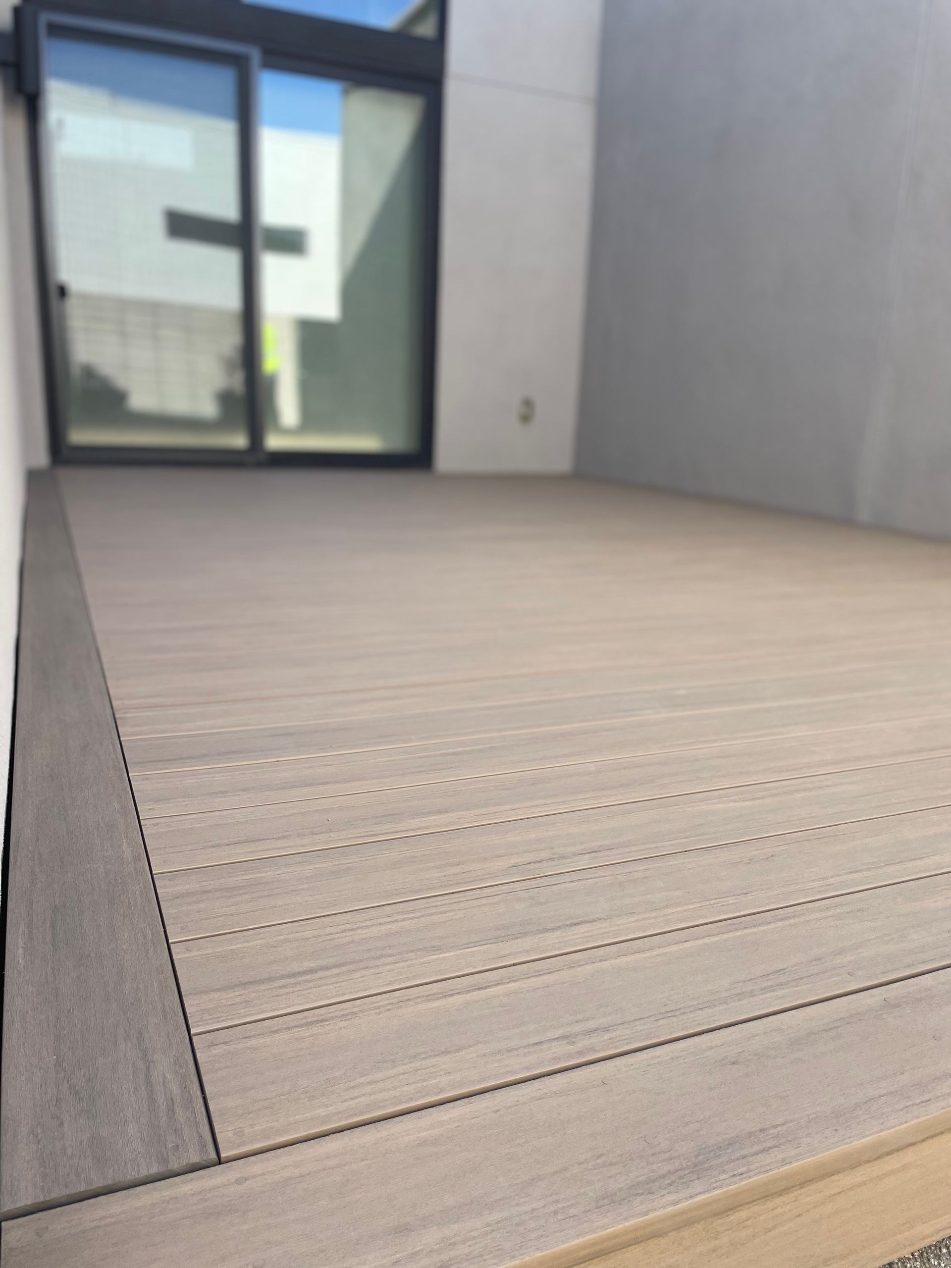 Modern wooden deck beside a gray wall and glass door leading to a patio.