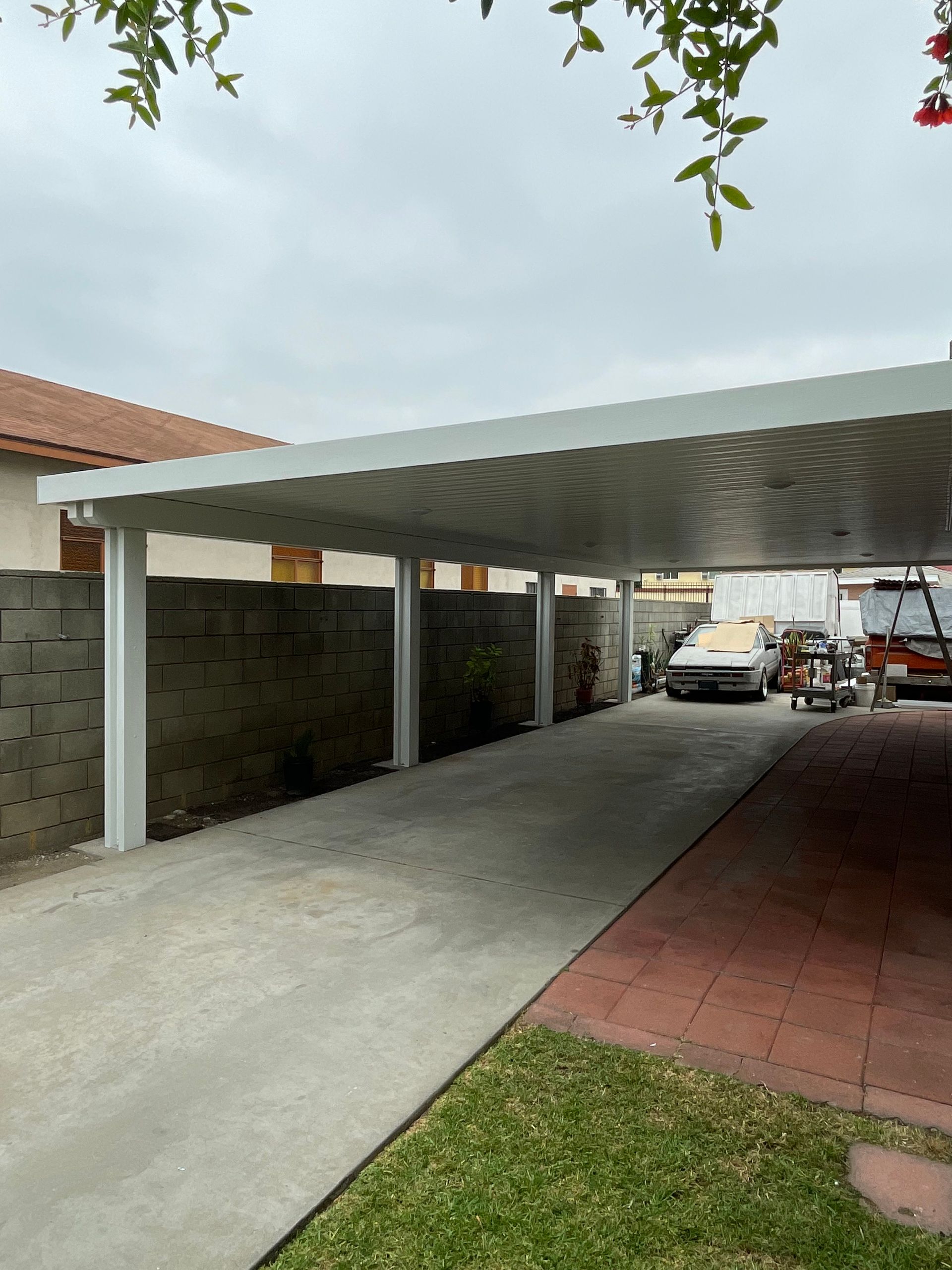 Covered driveway with white posts, concrete path, parked cars, and a small lawn beside a house