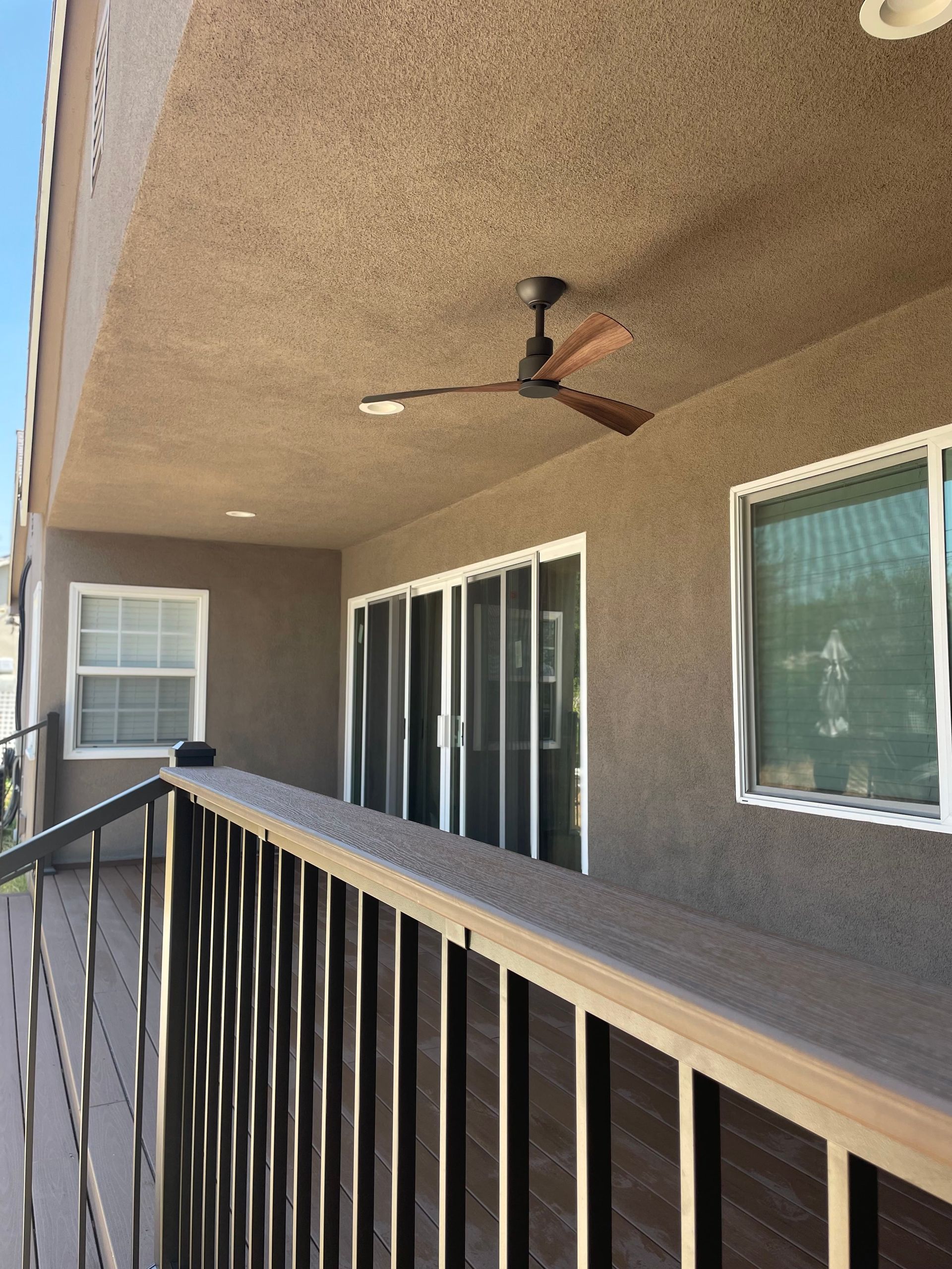 Covered patio with ceiling fan, black railing, and sliding glass doors beside tan stucco walls.