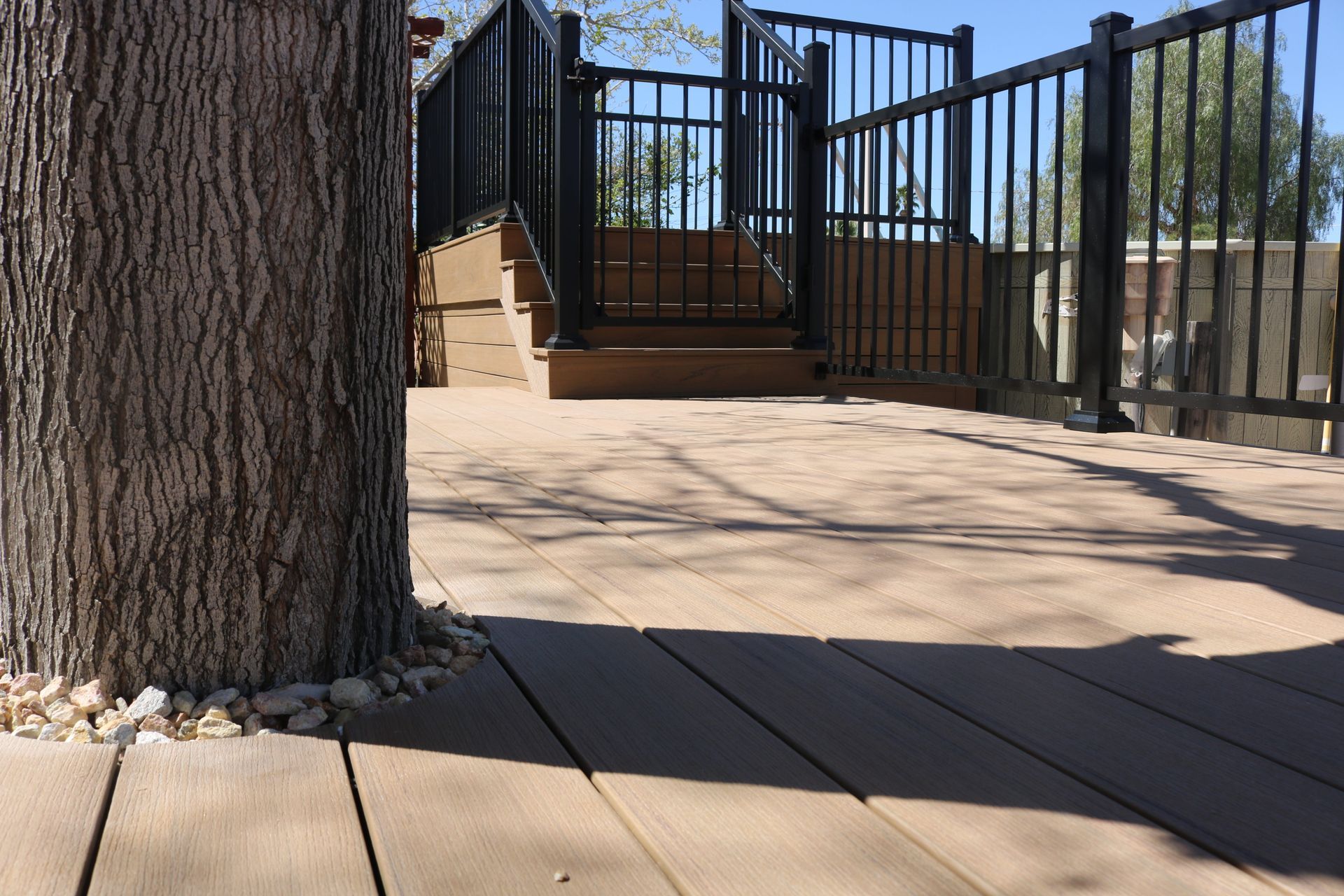 Wooden deck with black railing and a large tree trunk in the foreground, leading to steps and a gate.