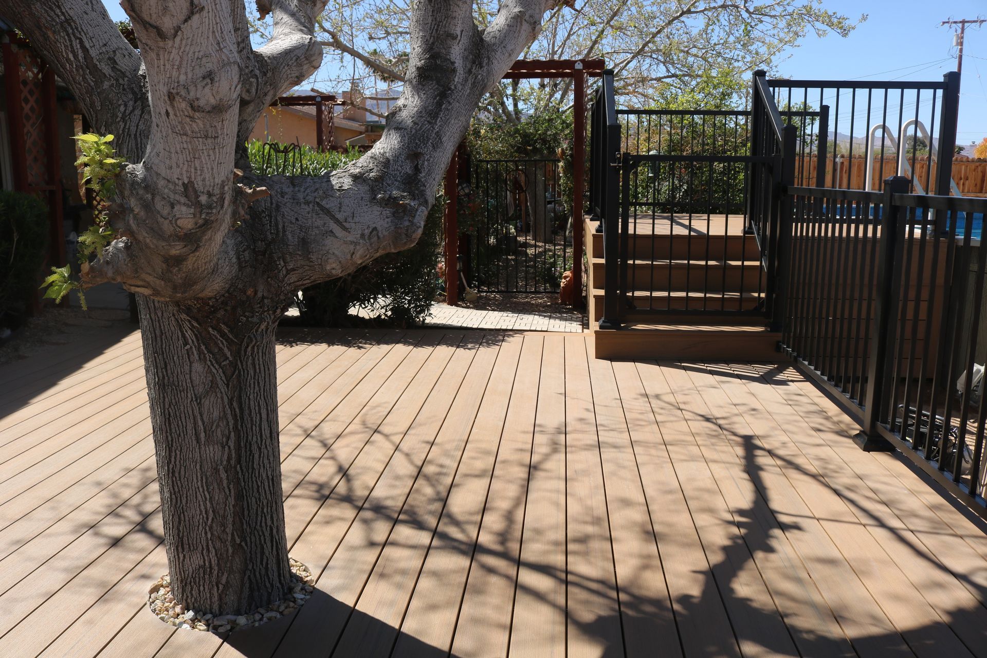 Wooden deck beside a tree and black metal fence, with steps leading to a gated entrance.