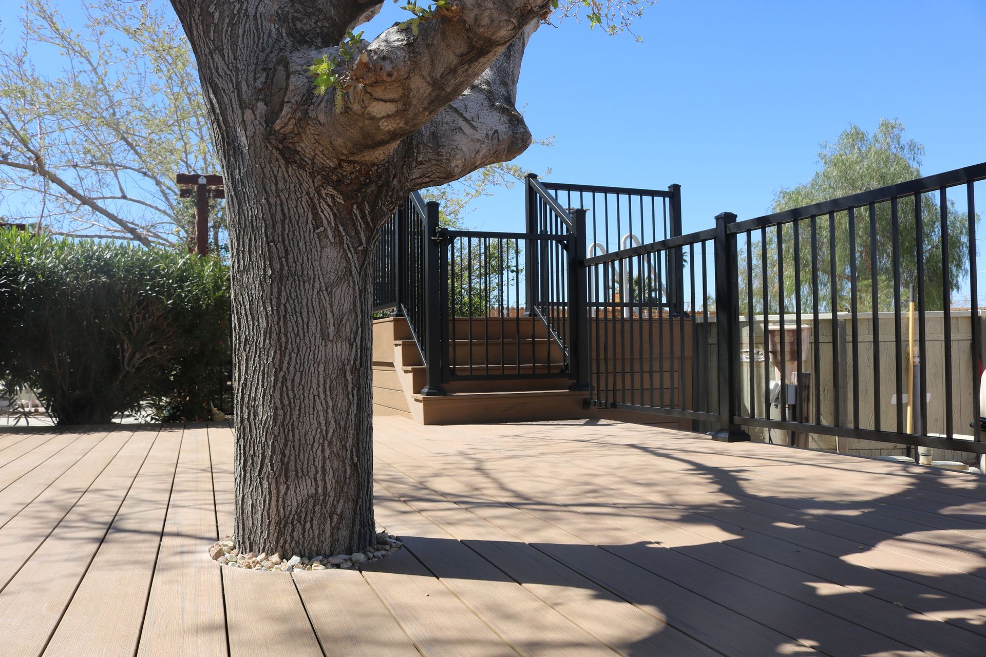 Tree-lined patio with brick pavers, a staircase, and black metal fencing under a clear blue sky