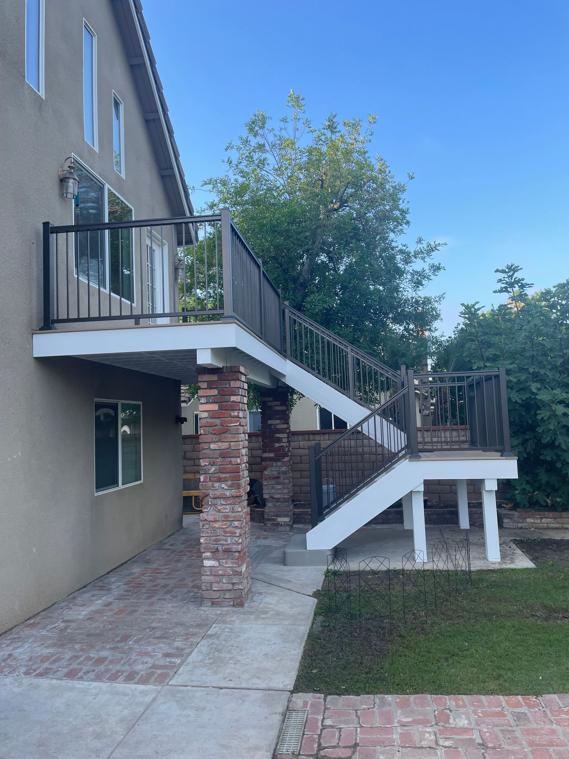 Outdoor staircase with white railings and brick supports beside a house, leading up to a deck amid trees.